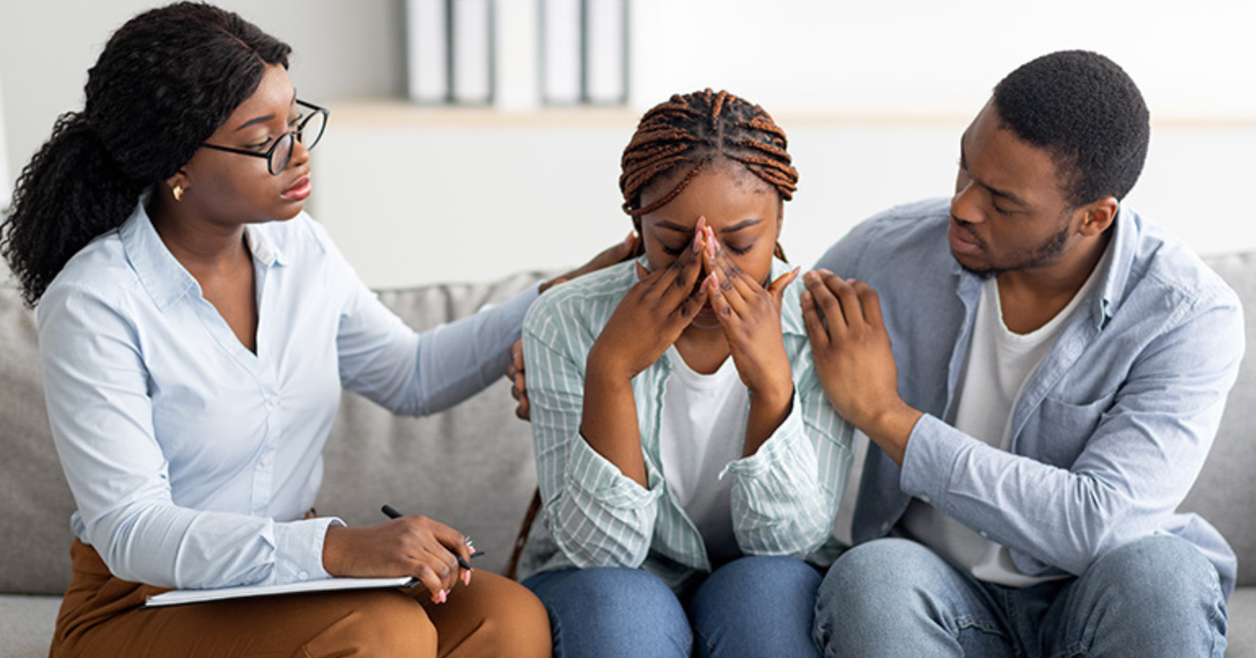 Three people sit on a couch; one woman is crying with her hands covering her face while the other two offer comfort and support, with one also holding a clipboard and pen.