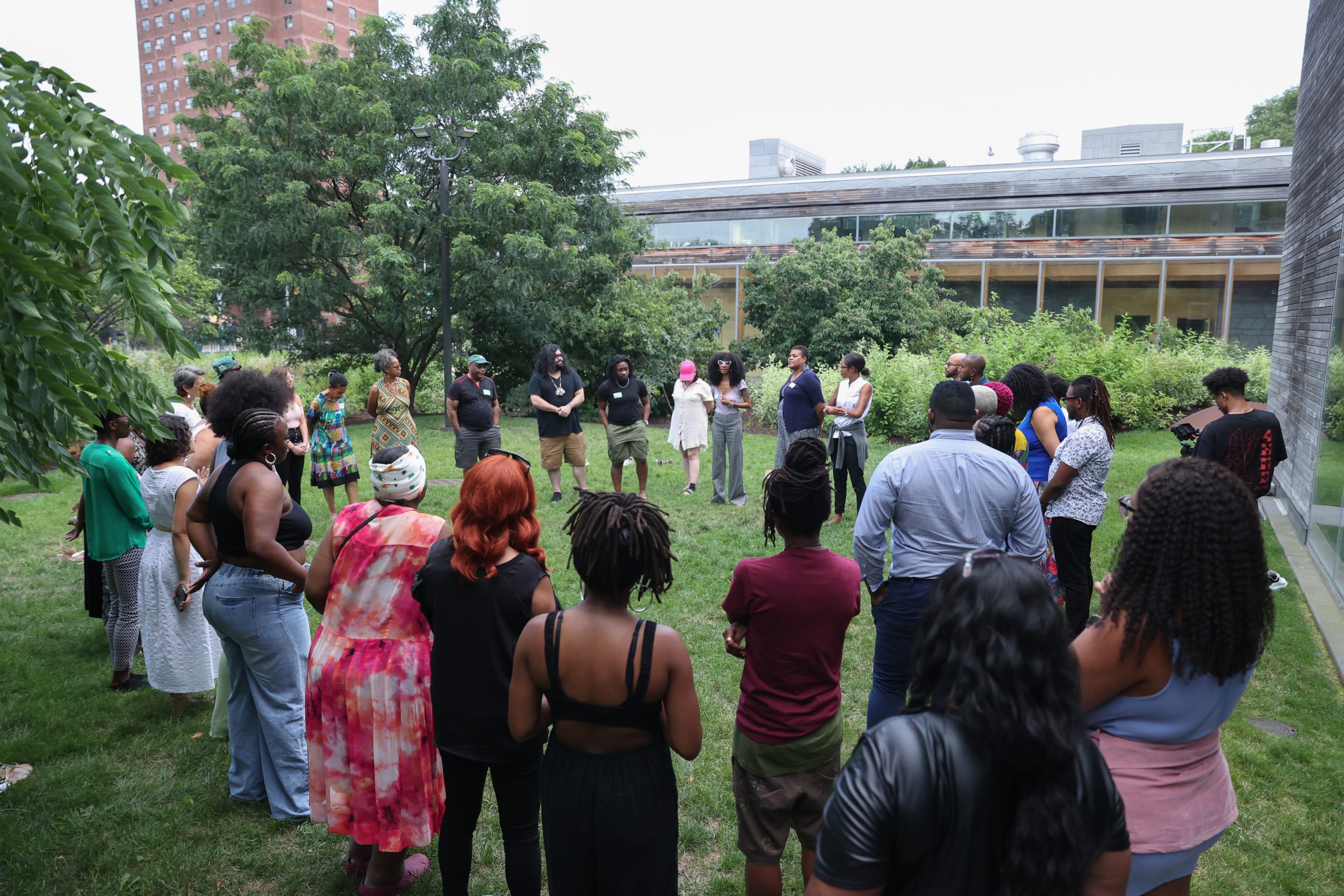 A group of people stand in a circle on a grassy area outdoors, engaged in conversation, with trees and buildings in the background.
