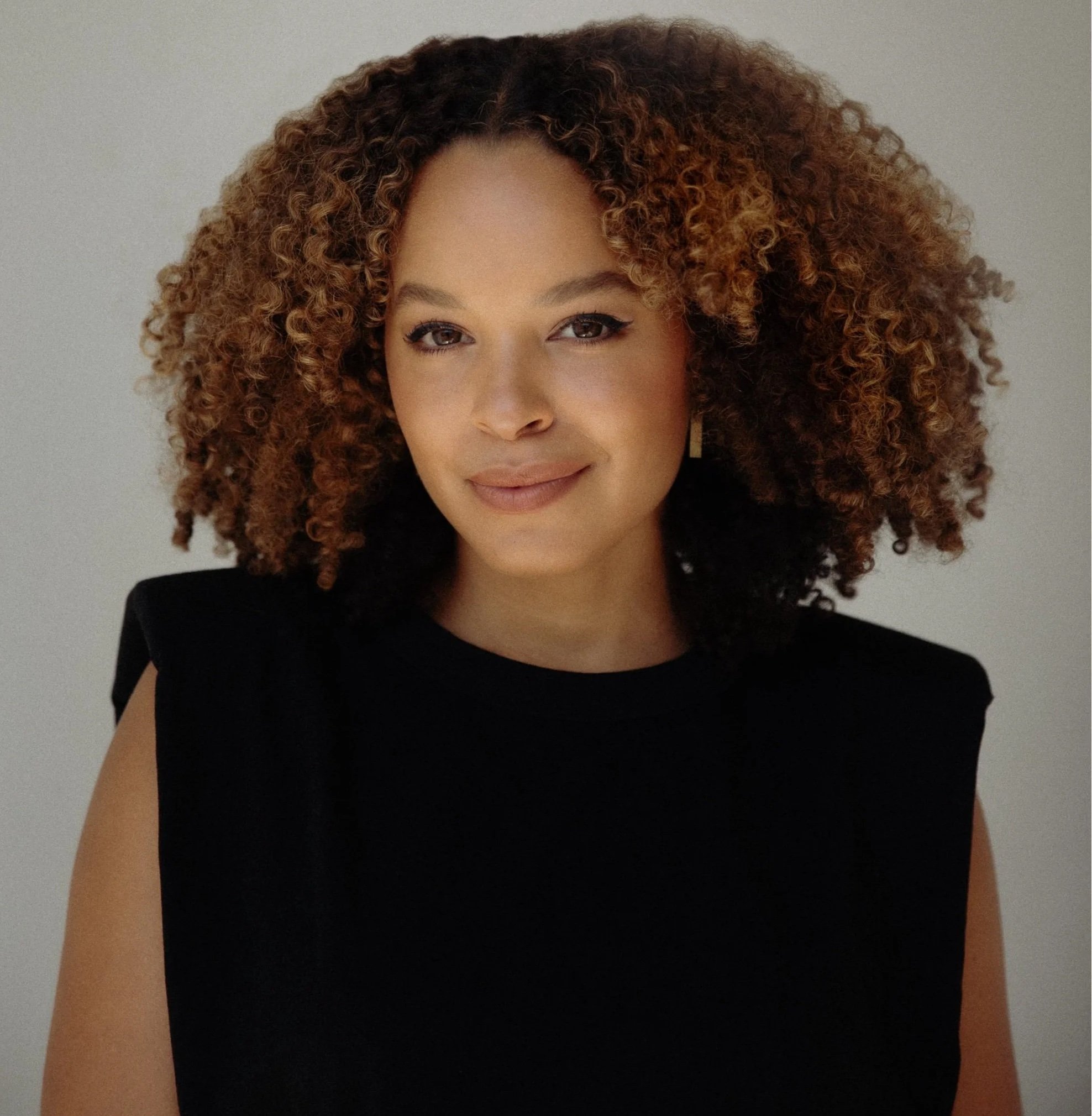 A woman with curly hair wearing a black sleeveless top stands against a plain light background, looking at the camera with a slight smile.