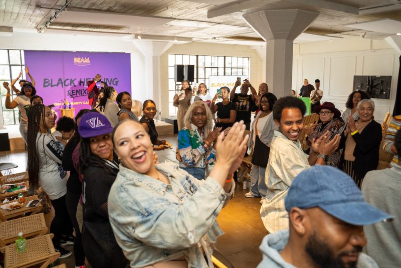 People clap and smile in a bright event space with “Black Healing Remix” displayed on a purple backdrop. Some are seated while others stand and interact in the lively atmosphere.