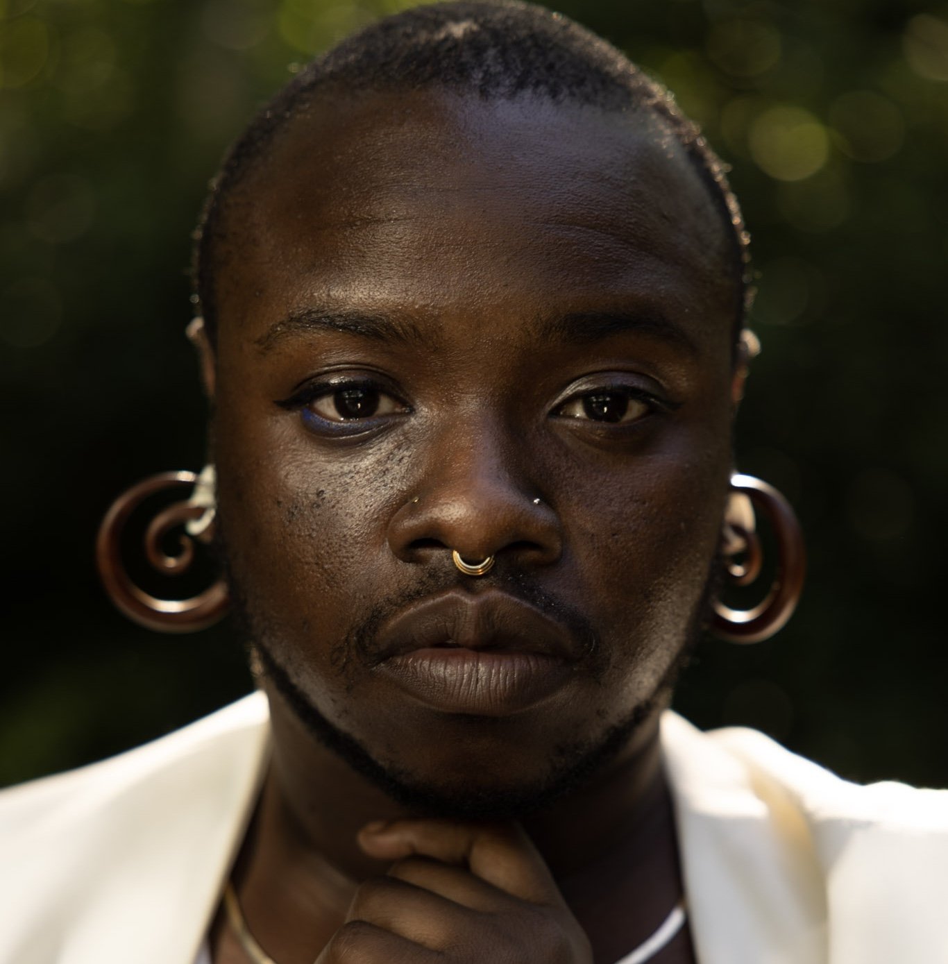 A person with gauged ears, a septum piercing, and short hair looks directly into the camera, resting their chin on their hand. The background is blurred greenery.