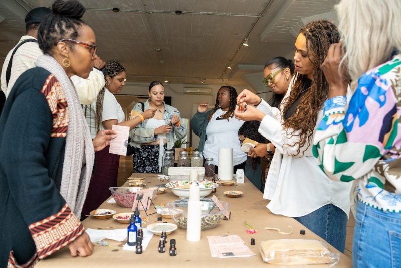 A group of people stand around a table with various items, examining small objects and papers in a well-lit indoor setting.