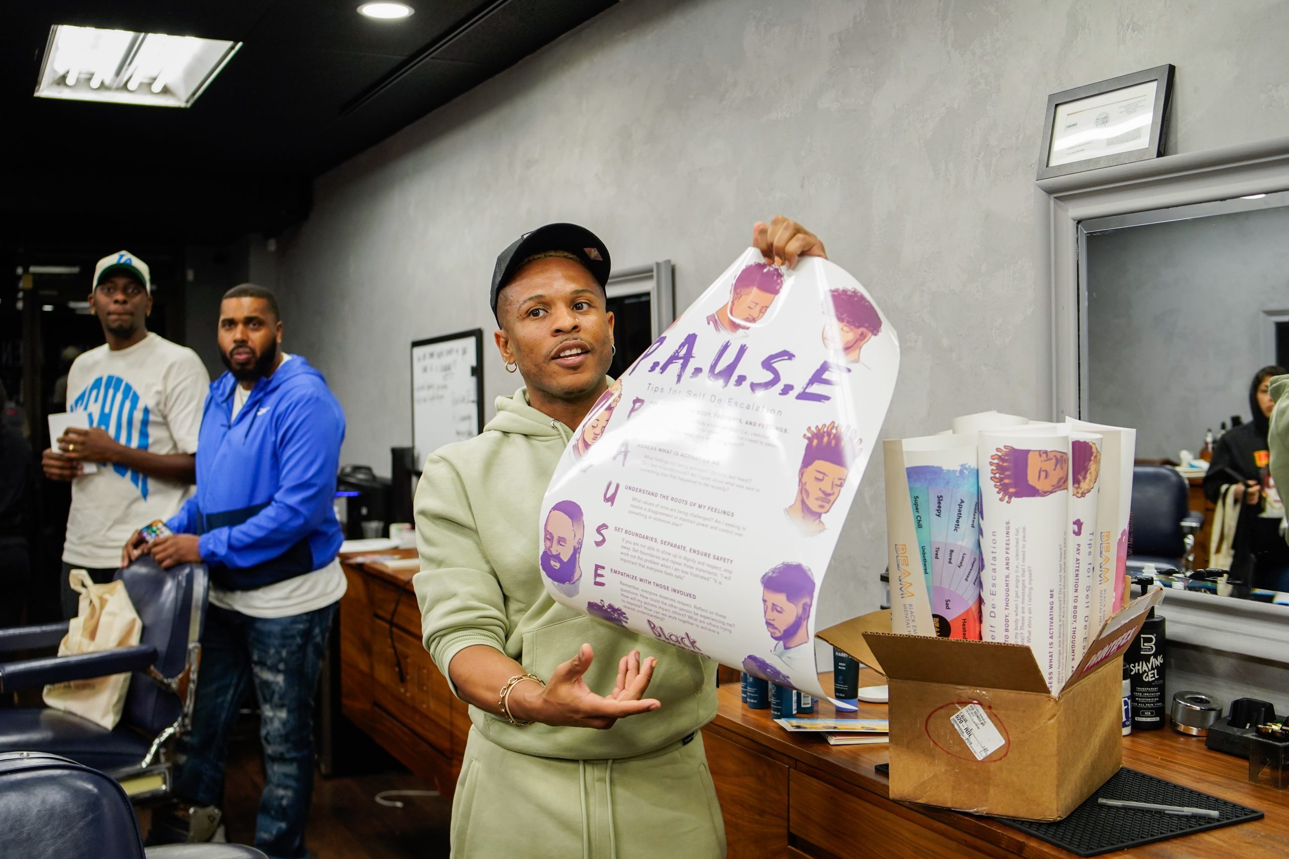 A person in a light green tracksuit holds up a colorful poster labeled "A.U.S.E" inside a barbershop, with two other people and mirrors visible in the background.