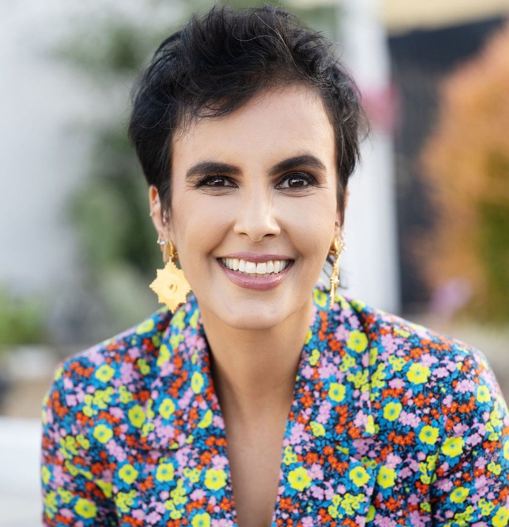 A woman with short dark hair smiles, wearing gold earrings and a colorful floral blazer, with a blurred outdoor background.