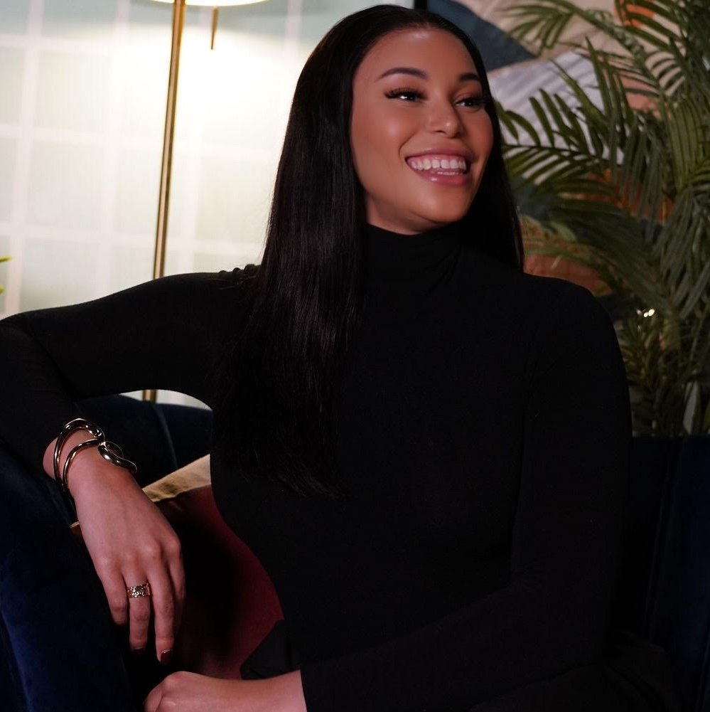 A woman in a black turtleneck sits on a dark sofa, smiling, with her hand resting on the armrest; a lamp and plants are visible in the background.
