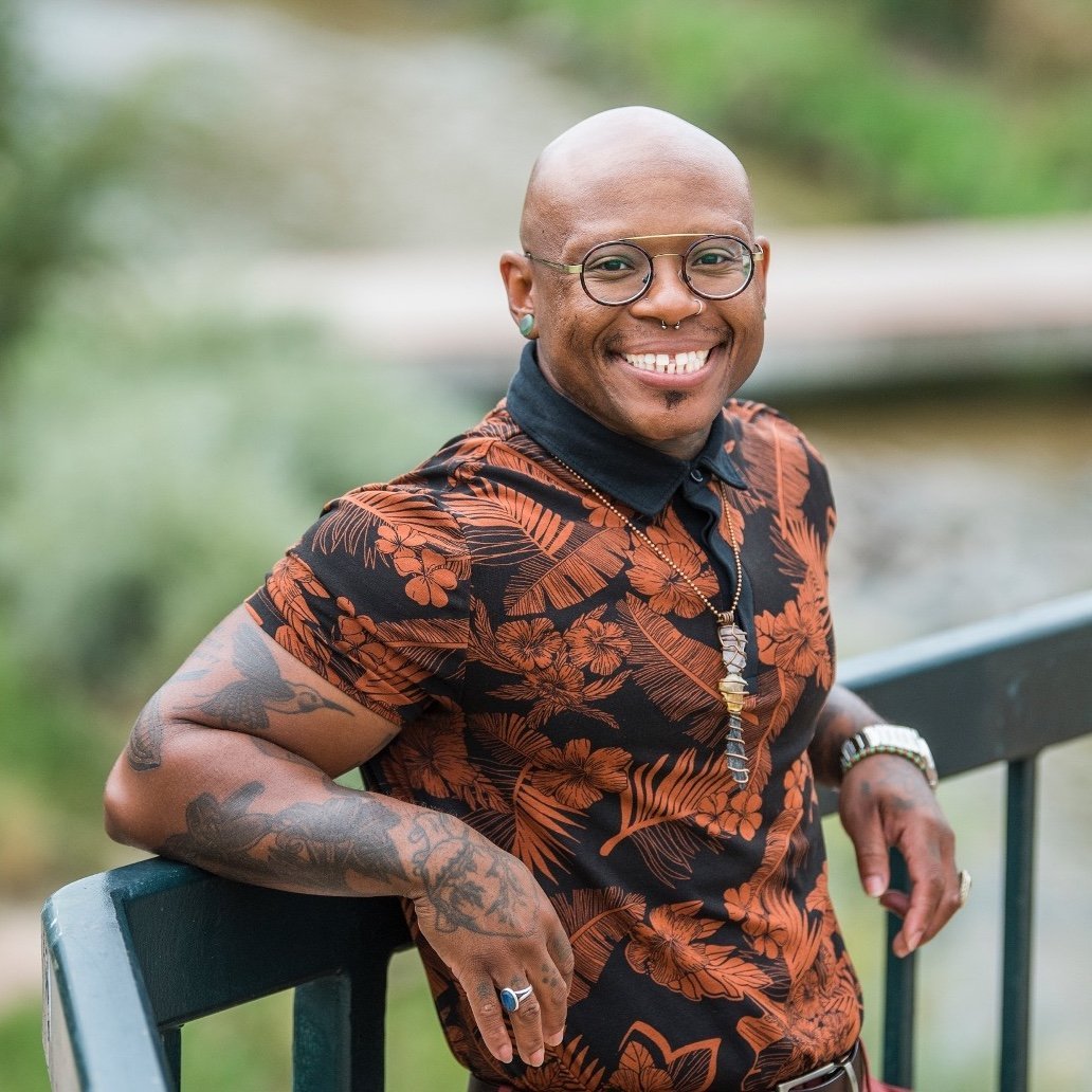 A person with glasses, tattoos, and jewelry, wearing a floral shirt, leans on a railing outdoors and smiles at the camera.