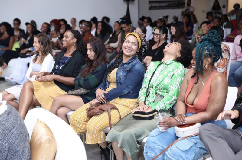 A diverse group of people sit in rows at an indoor event, with many smiling and laughing, dressed in colorful, casual clothing.