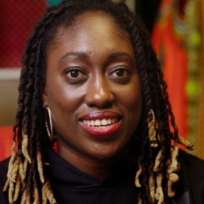 A woman with long, dark, twisted hair and blonde tips smiles at the camera, wearing hoop earrings and red lipstick, with a colorful background.