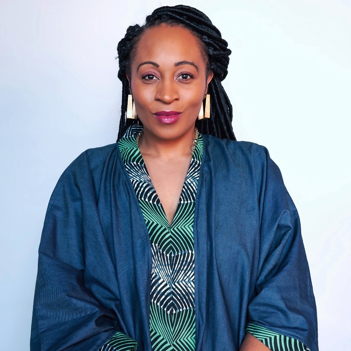 A woman with long braided hair wears geometric-patterned green and black clothing with a navy blue outer layer, accessorized with large rectangular earrings, against a plain light background.