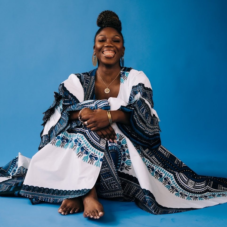 Woman sits barefoot on the floor against a blue background, smiling broadly and wearing a white and blue patterned robe with large hoop earrings and jewelry.