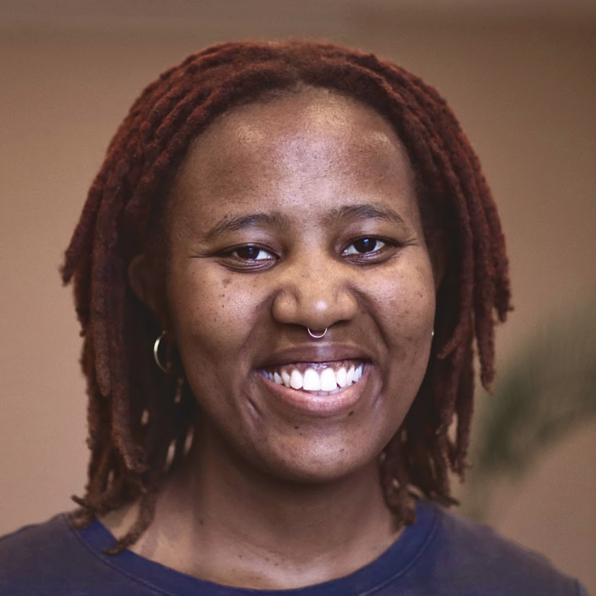 A person with short, reddish-brown dreadlocks, a septum piercing, and hoop earrings smiles at the camera against a neutral background.