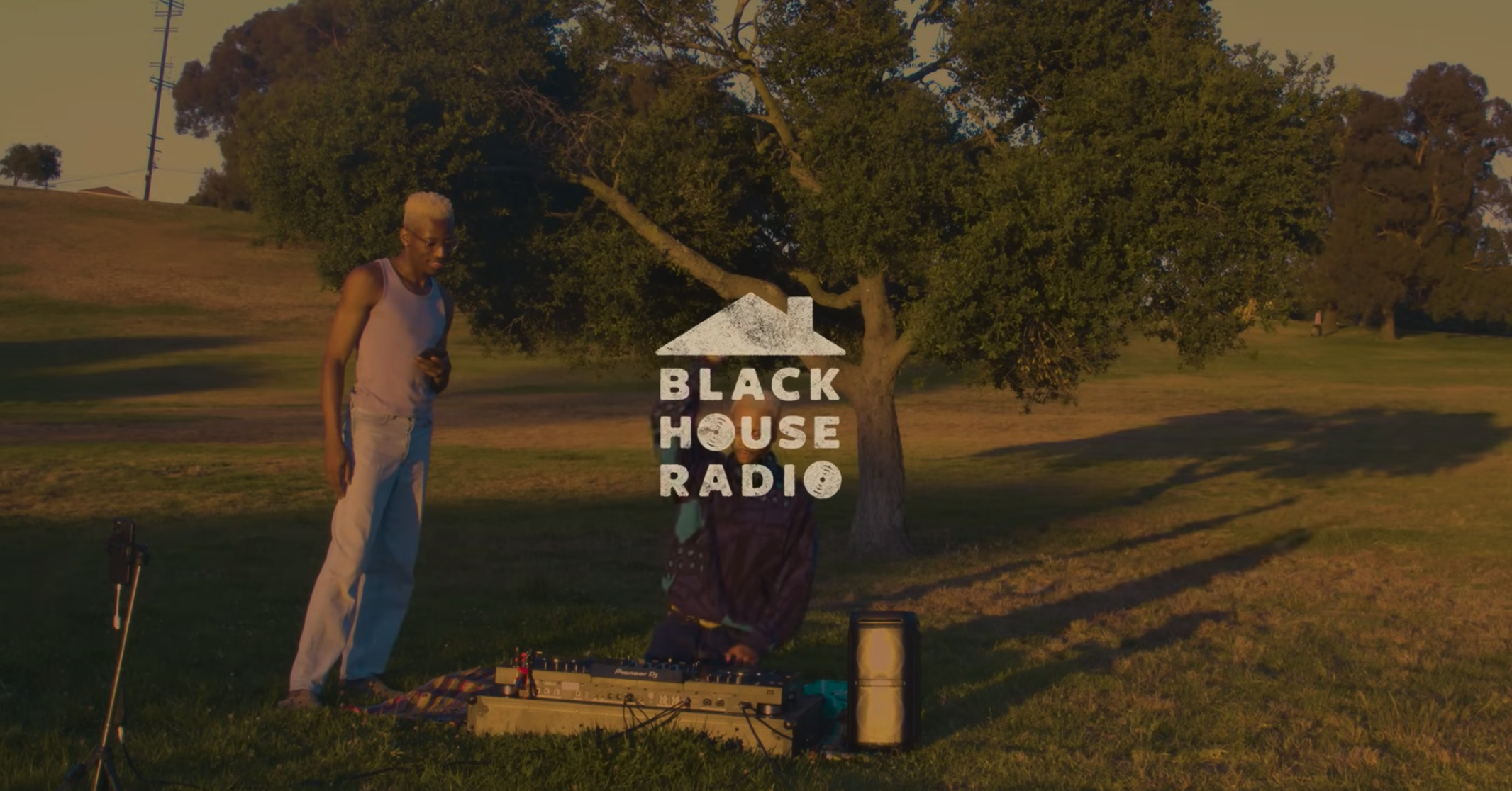 Two people stand in a grassy park near music equipment at sunset; the words "BLACK HOUSE RADIO" with a house icon are overlaid in the center.