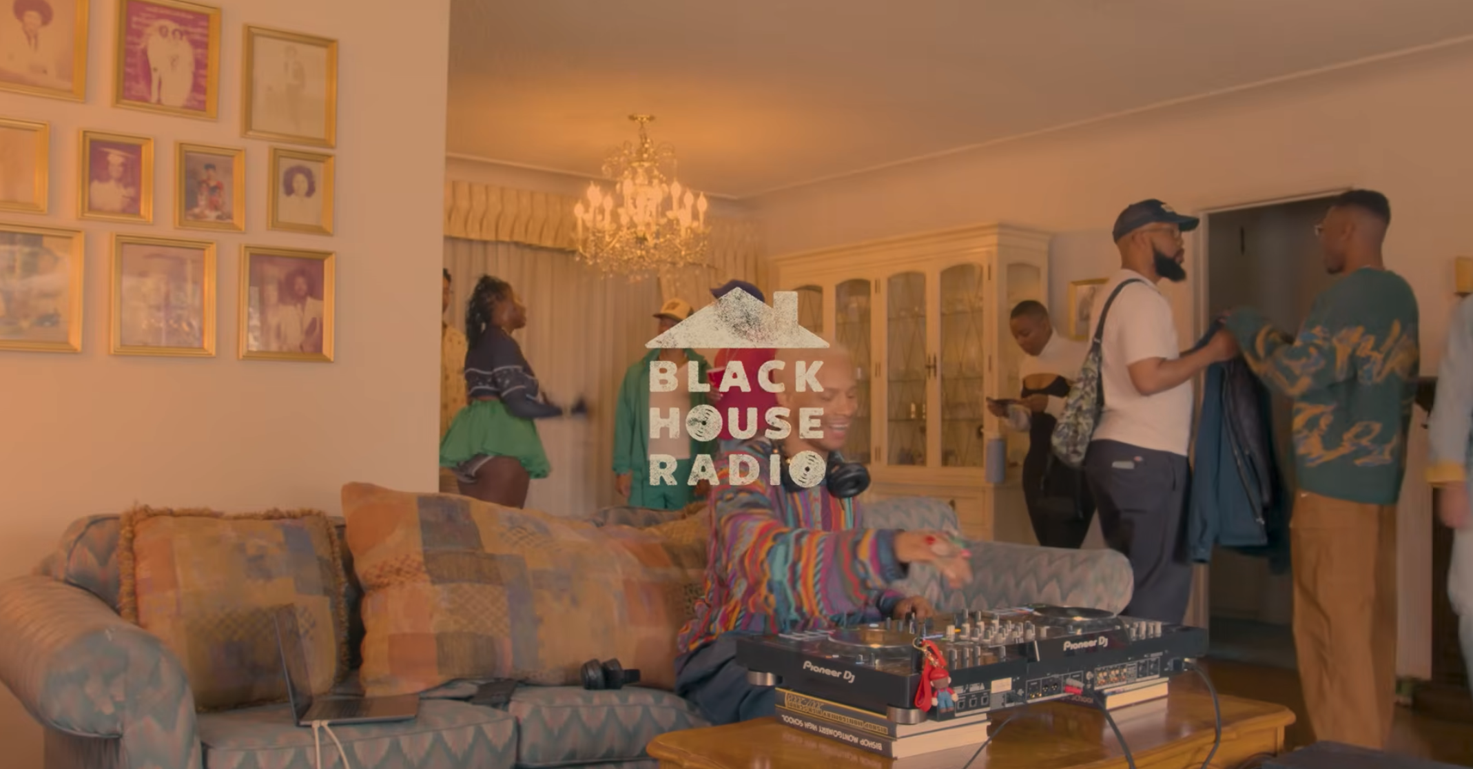A group of people gather in a living room; one person operates DJ equipment on a table while others talk and move around. The words "Black House Radio" are displayed in the center.