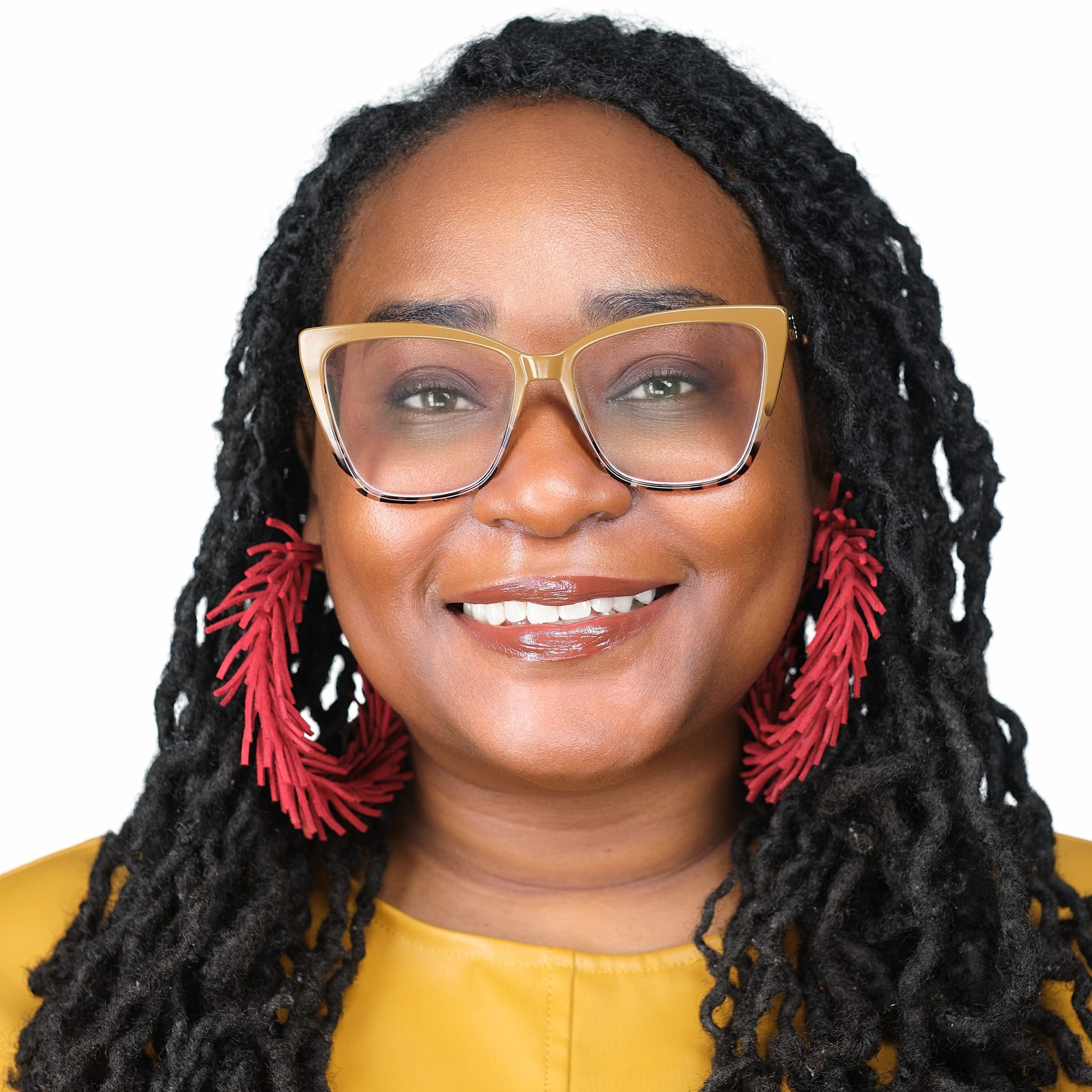 A woman with long black locs, large glasses, and red statement earrings smiles at the camera. She is wearing a mustard yellow top. The background is white.