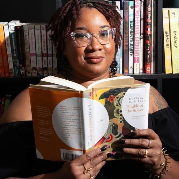 A person with glasses holds and reads "Parable of the Sower" by Octavia E. Butler in front of a bookshelf filled with various books.