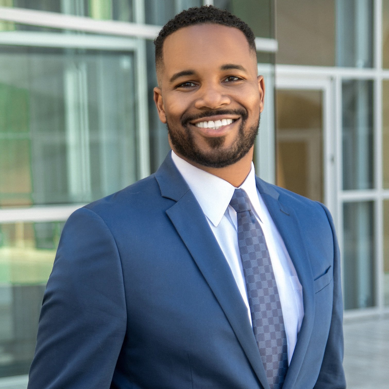 A man in a blue suit and tie stands outside a modern glass building, smiling at the camera.