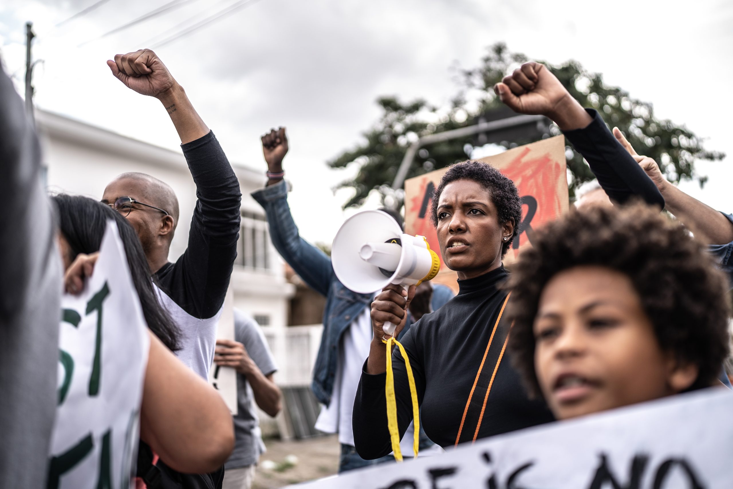A group of people protest outdoors, raising fists; a woman holds a megaphone and speaks, with a child in the foreground and signs visible in the background.