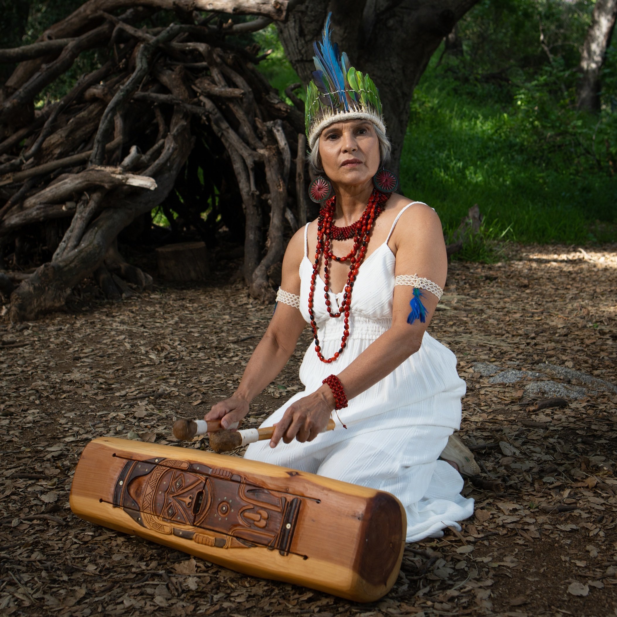 A woman in a white dress and feathered headdress sits on the ground in a wooded area, playing a carved wooden drum with mallets.